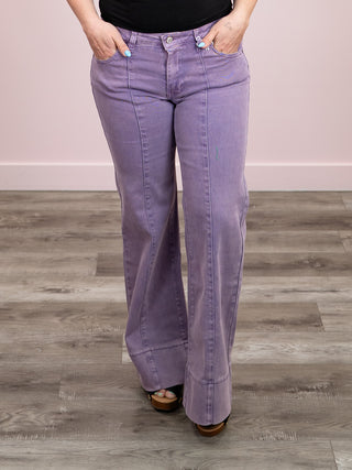 A person wearing lavender-purple wide-leg jeans with a black top and brown sandals, standing on a gray wood floor against a light pink wall.