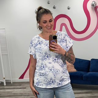 A woman takes a mirror selfie in a boutique showing a white short-sleeve t-shirt with a blue floral pattern, paired with light-wash wide-leg jeans and brown slide sandals. The background features a gray wood-look floor, a blue velvet sofa, and a white wall with a pink wavy graphic.